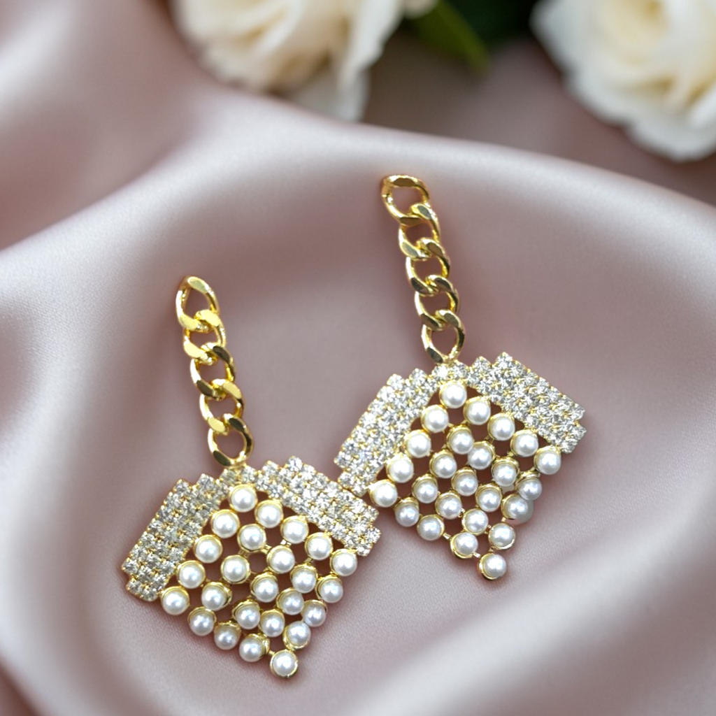 A pair of gold-tone drop earrings with square clusters of white faux pearls and clear rhinestones, resting on a soft pink satin fabric with blurred white roses in the background.